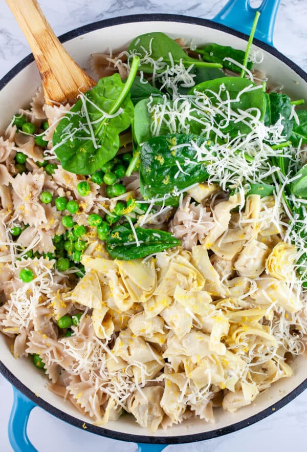 Cooked pasta, marinated artichoke hearts, peas, fresh spinach, and grated Parmesan cheese in skillet with wooden spoon.