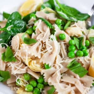 Spring vegetable pasta garnished with Parmesan cheese and lemon wedges in white bowl with fork.