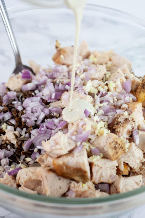 Creamy dressing poured into glass bowl with diced chicken, wild rice, and minced garlic and shallots.