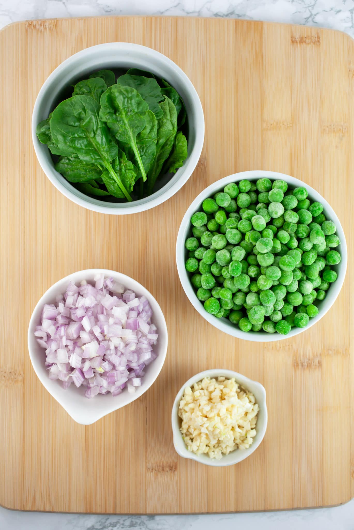 Minced garlic, shallots, frozen peas, and spinach in small white bowls on wooden cutting board.