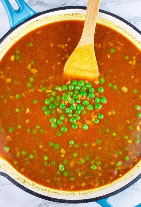 Chicken broth, tomato sauce, and peas in skillet with wooden spatula.