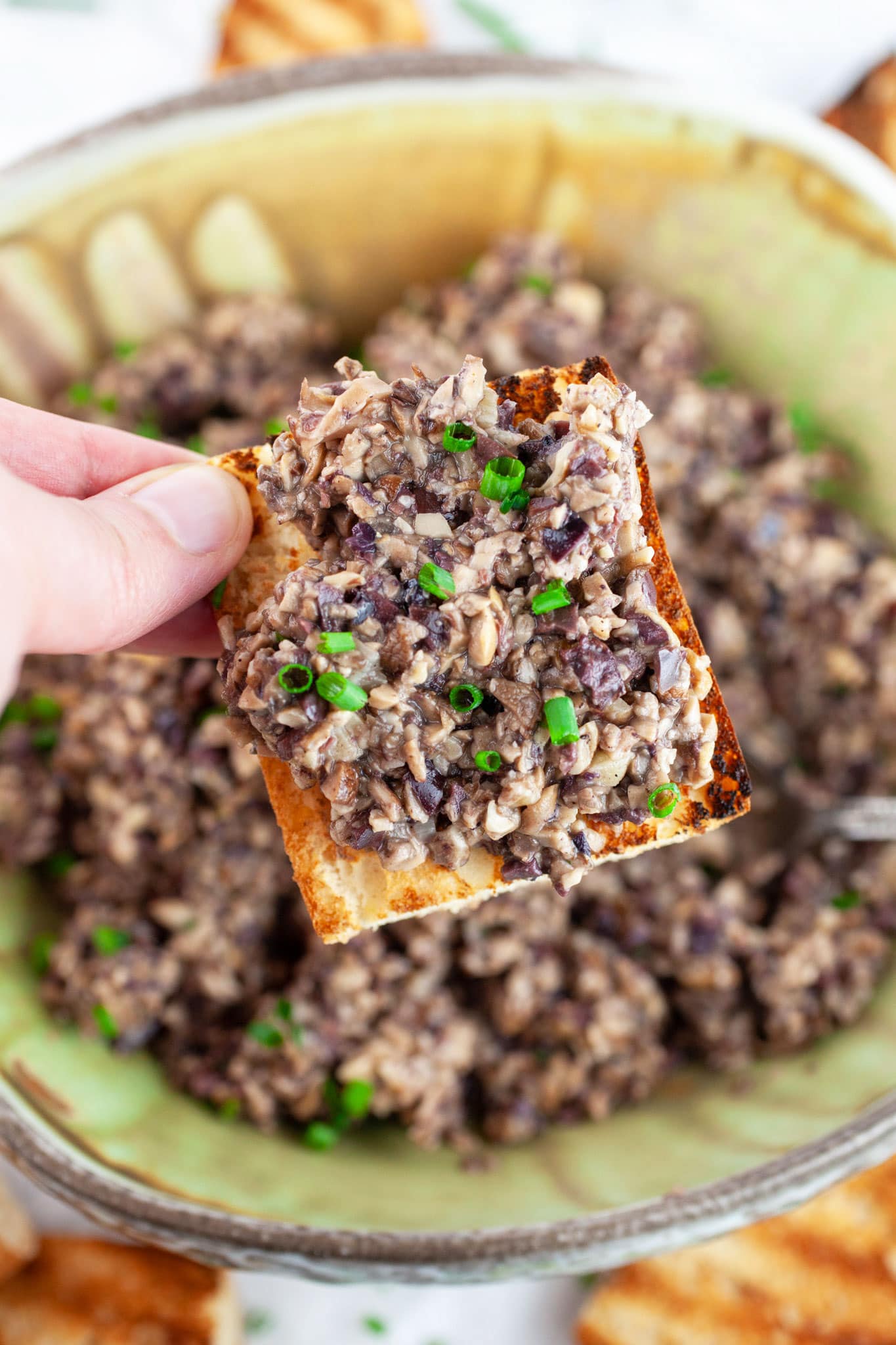 Hand holding mushroom tapenade on grilled baguette over ceramic bowl.