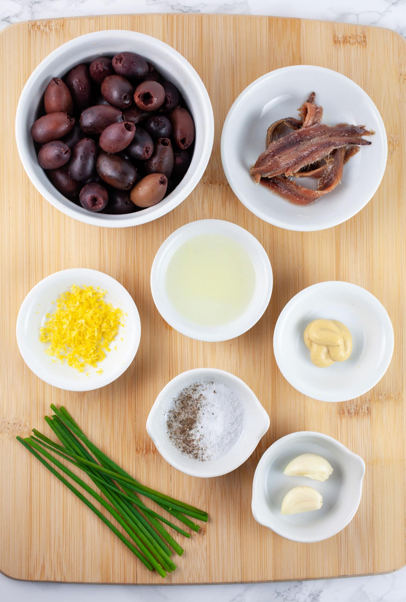 Garlic cloves, Dijon mustard, kosher salt and pepper, lemon zest and juice, anchovy filets, Kalamata olives, and fresh chives on wooden cutting board.