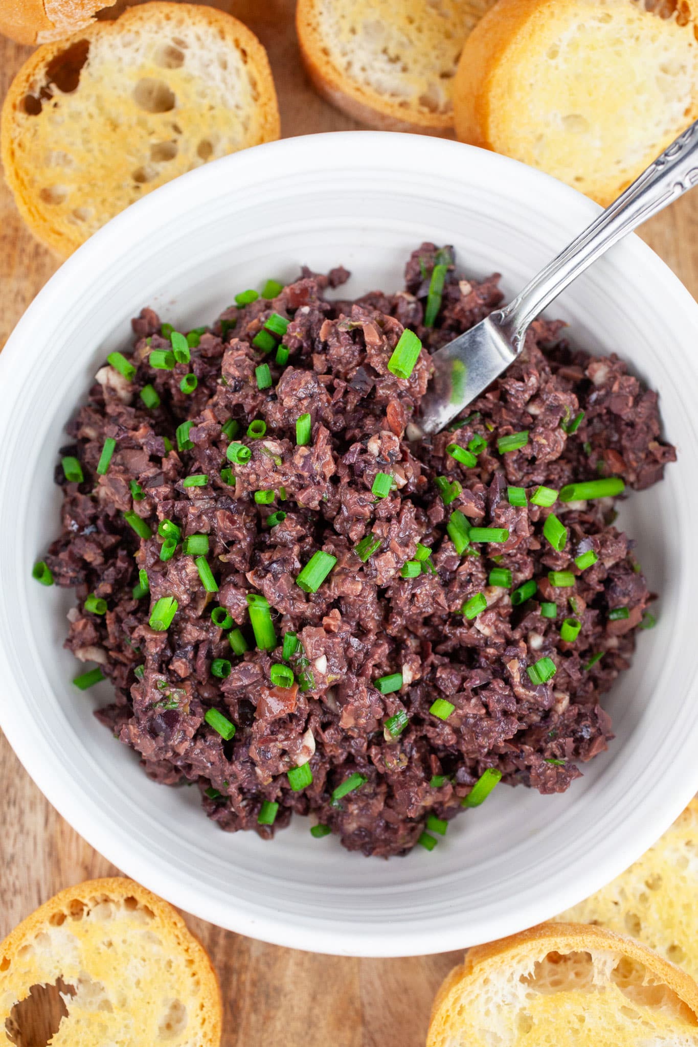 Kalamata olive tapenade garnished with fresh chives in white bowl with knife surrounded by crostini on wooden serving tray.