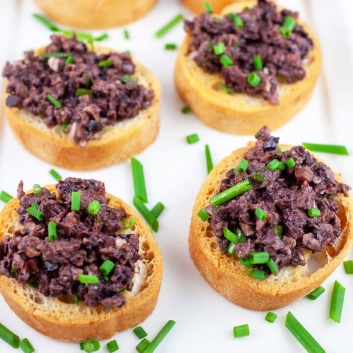 Kalamata olive tapenade on crostini garnished with fresh chives on white serving platter.