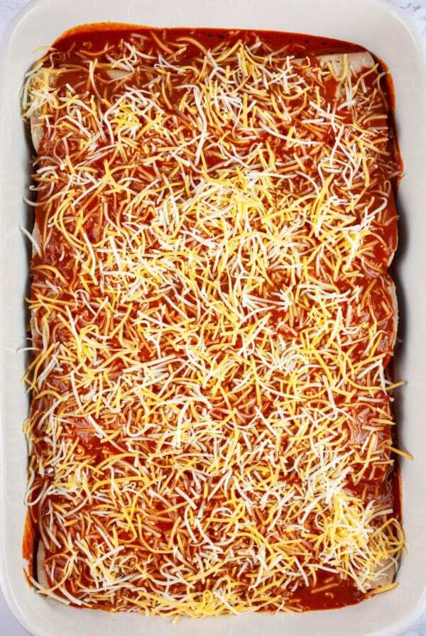 Filled tortillas covered with red enchilada sauce and shredded cheese in casserole dish.