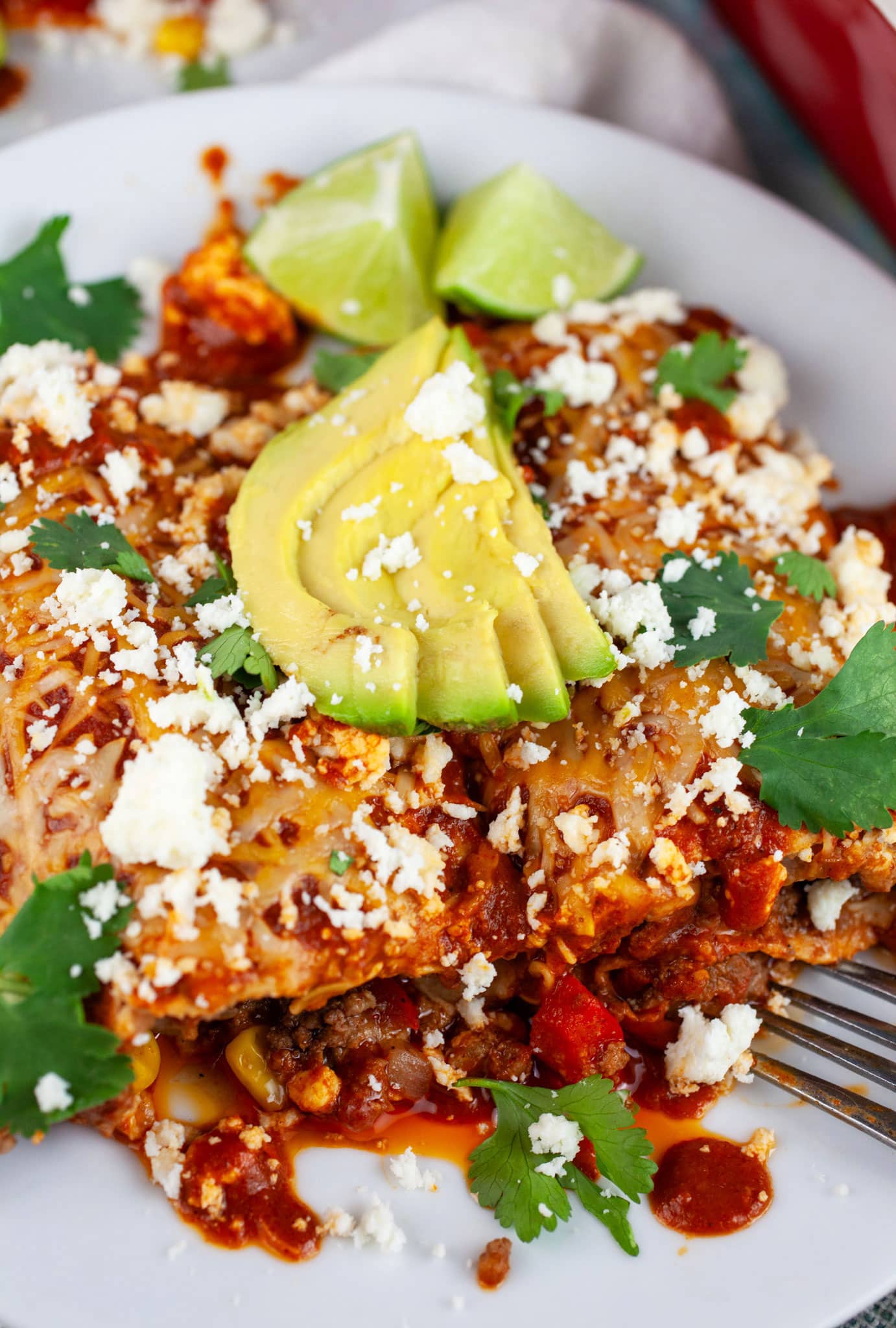 Ground beef enchiladas on white plate with fork garnished with Queso fresco, cilantro, sliced avocadoes, and limes.
