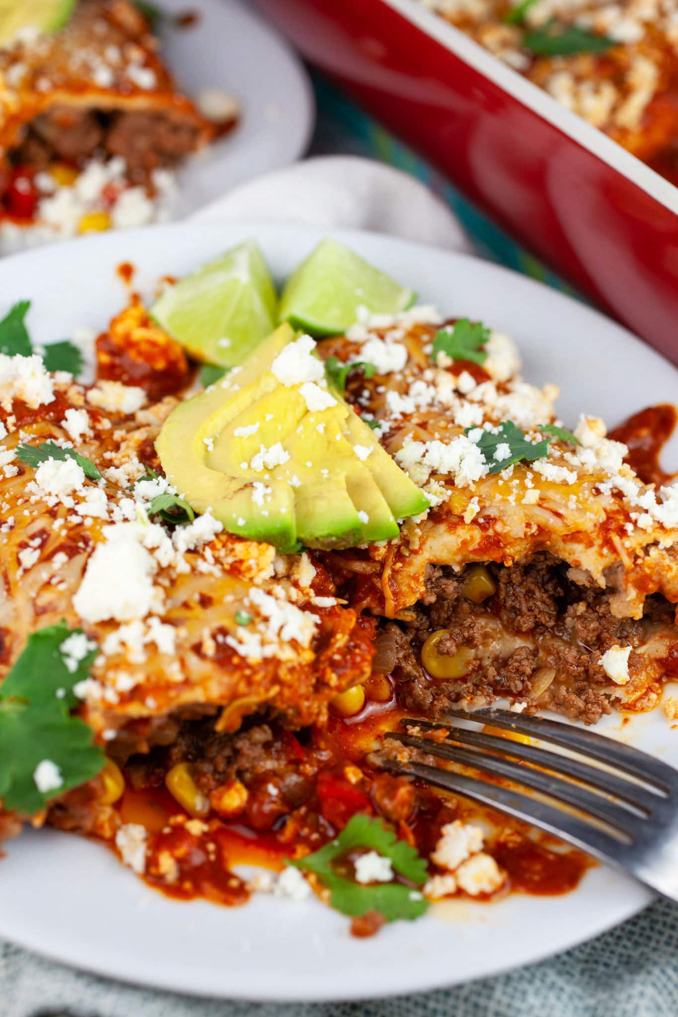 Ground beef enchiladas with Queso fresco, sliced avocadoes, cilantro, and lime wedges on white plate with fork.