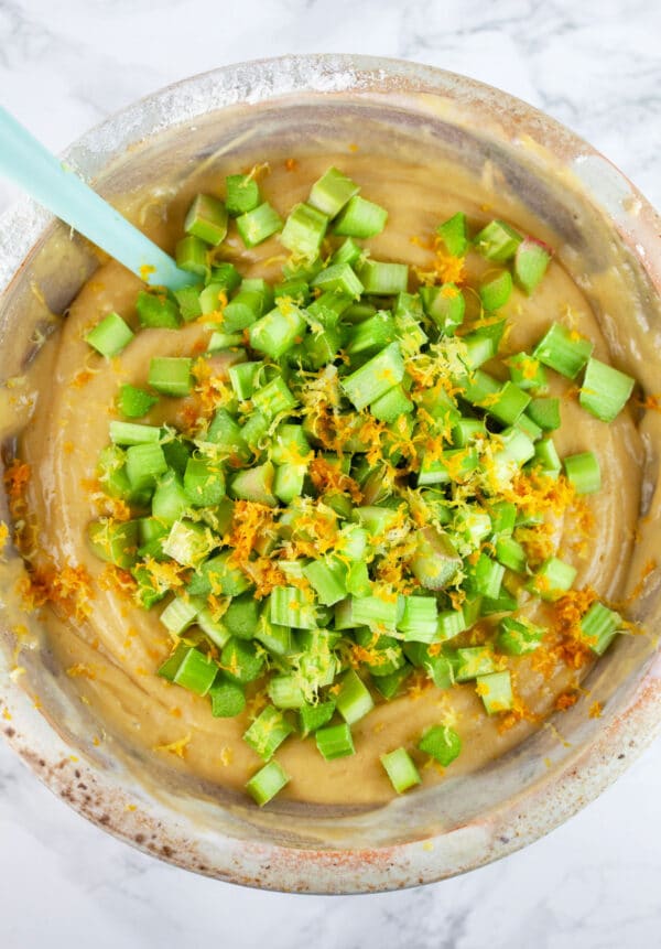 Diced rhubarb and orange and lemon zest added to batter in ceramic bowl with blue spatula.