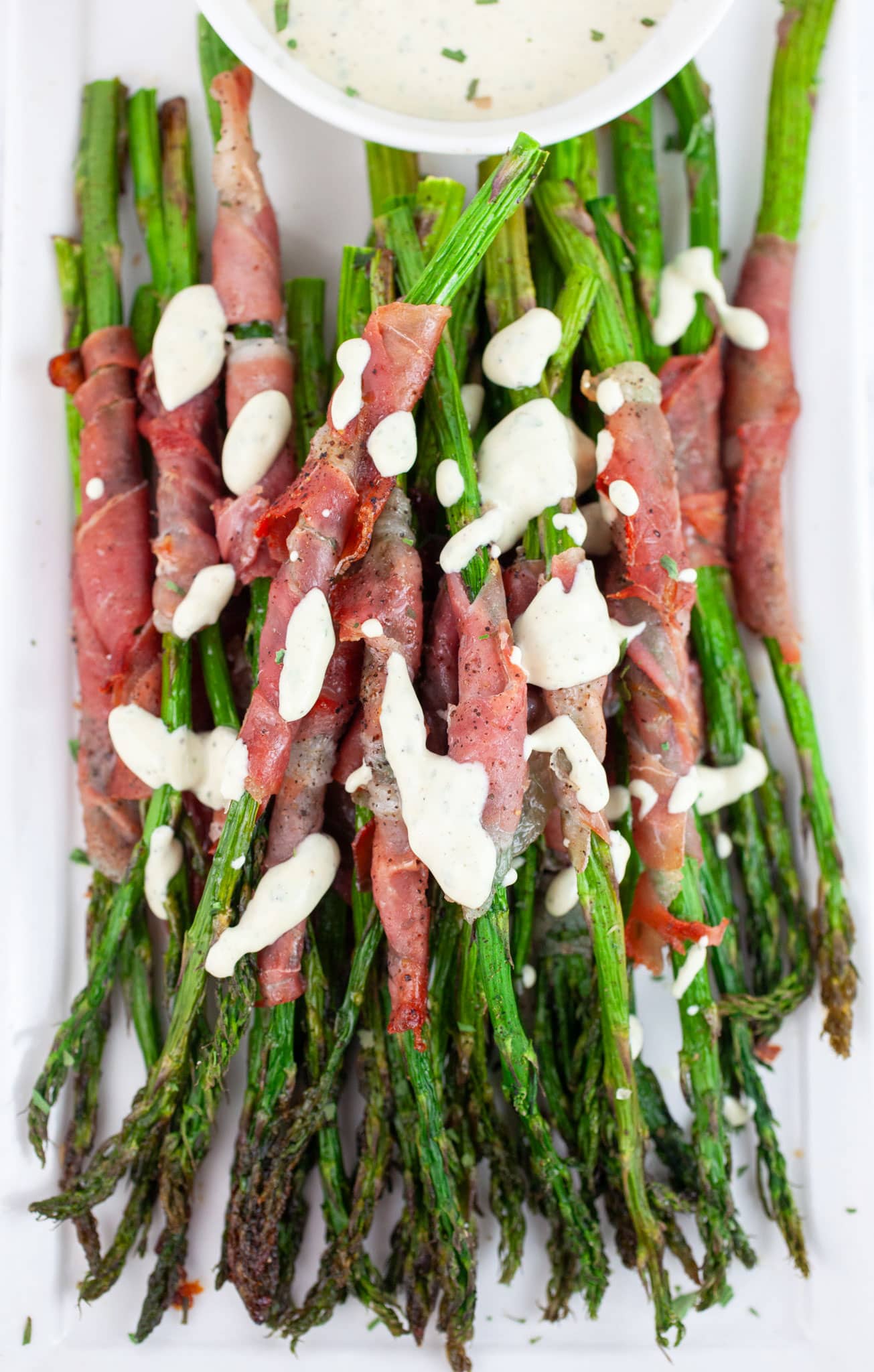 Roasted prosciutto wrapped asparagus drizzled with tarragon sauce on white serving platter.