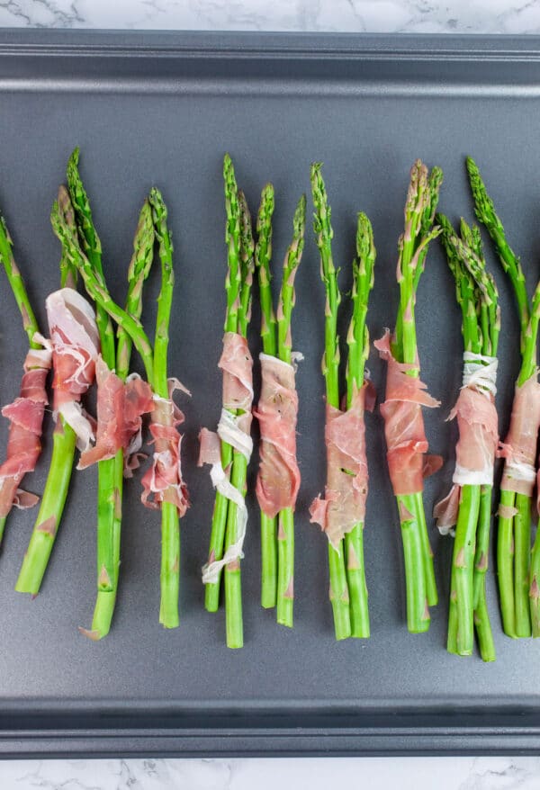 Uncooked asparagus with prosciutto on baking sheet.