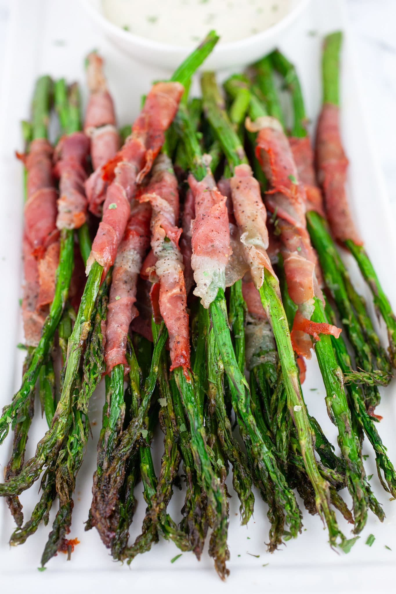 Roasted prosciutto wrapped asparagus on white serving platter with small bowl of creamy dipping sauce.