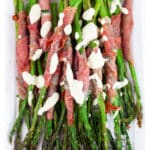 Roasted prosciutto wrapped asparagus drizzled with tarragon sauce on white platter.