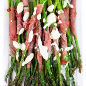 Roasted prosciutto wrapped asparagus drizzled with tarragon sauce on white platter.