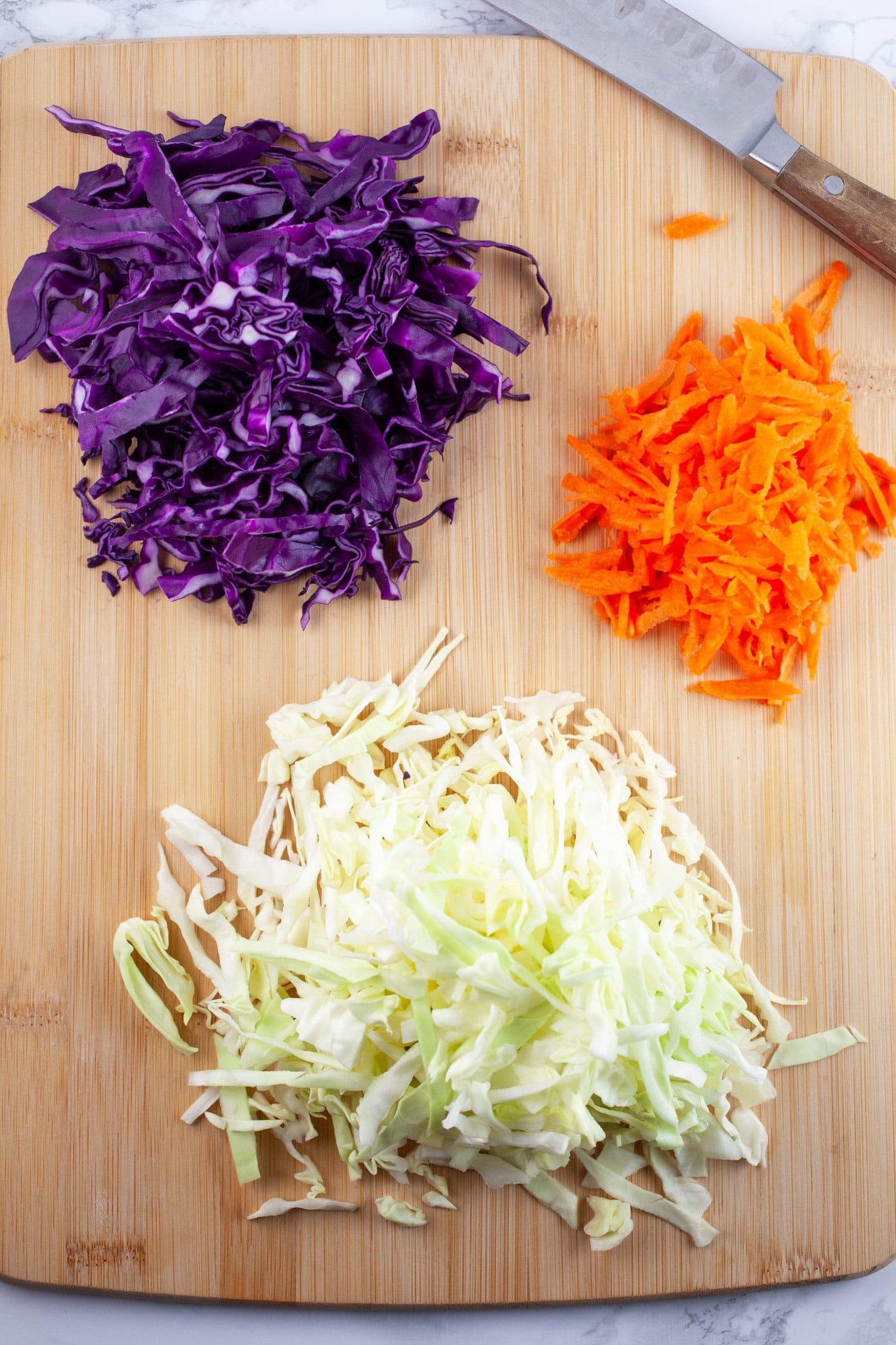 Shredded green and red cabbage and carrots on wooden cutting board with knife.