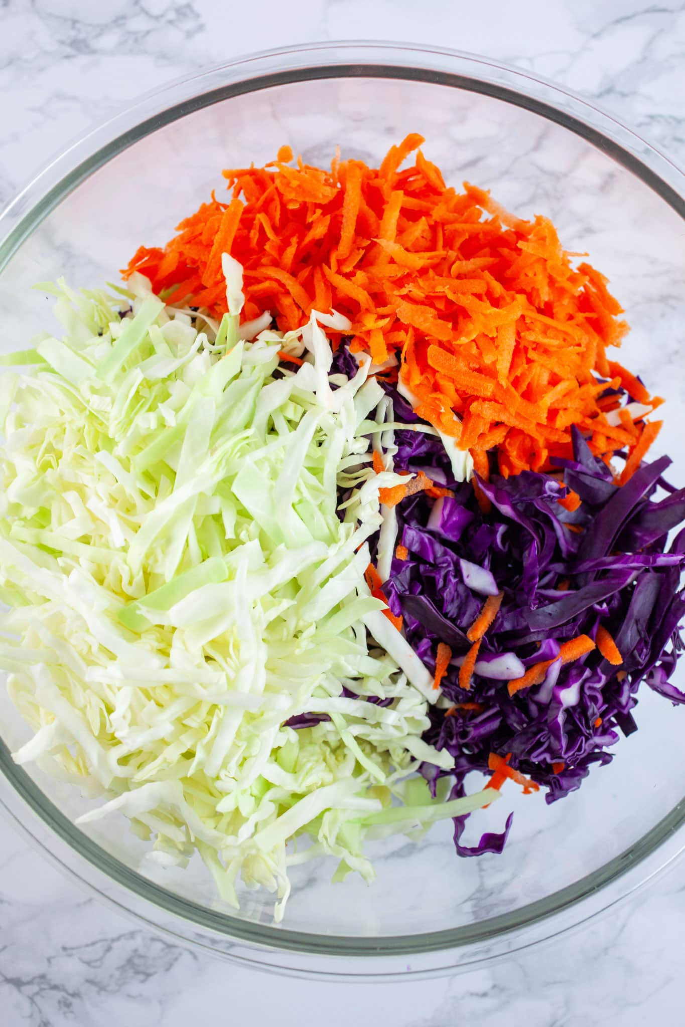 Shredded green and red cabbage and carrots in large glass bowl.