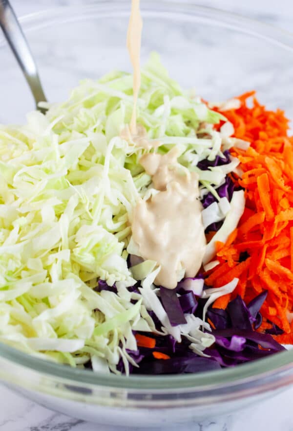 Miso ginger dressing poured onto shredded cabbage and carrots in glass mixing bowl with spoon.