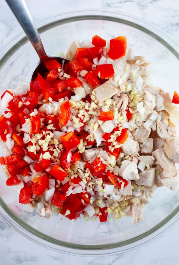 Diced chicken, garlic and shallots, and red bell peppers in glass mixing bowl with spoon.