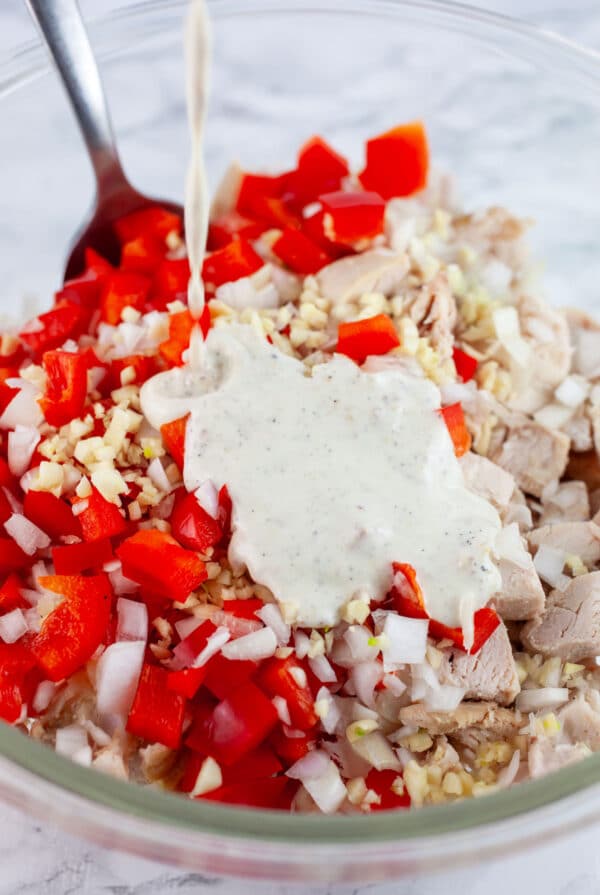 Lemon mayo dressing poured onto chicken salad ingredients in glass bowl with spoon.