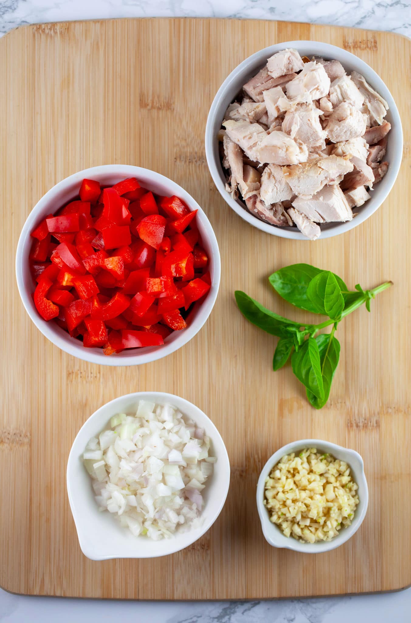 Minced garlic, shallots, red bell peppers, rotisserie chicken, and fresh basil on wooden cutting board.
