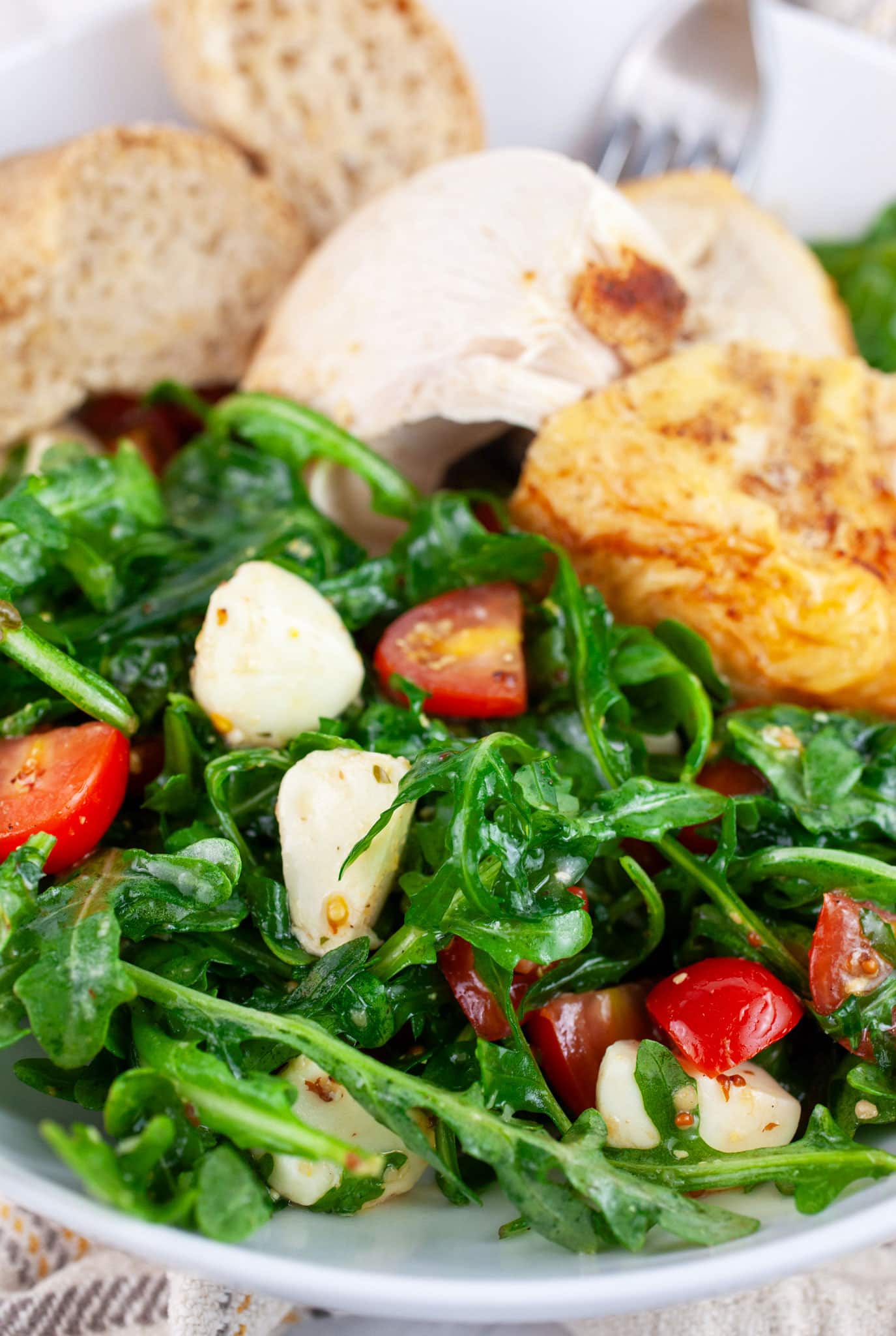 Arugula tomato mozzarella salad with roasted chicken and toasted baguette in white bowl with fork.