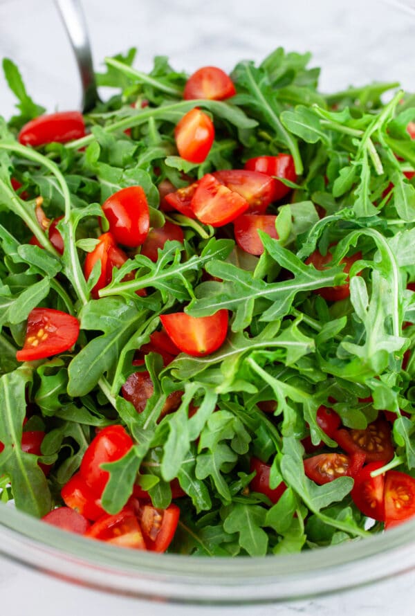 Arugula and chopped grape tomatoes in glass bowl with spoon.