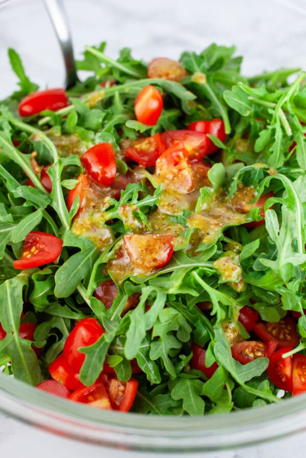 Arugula, chopped grape tomatoes, and vinaigrette in glass bowl with spoon.
