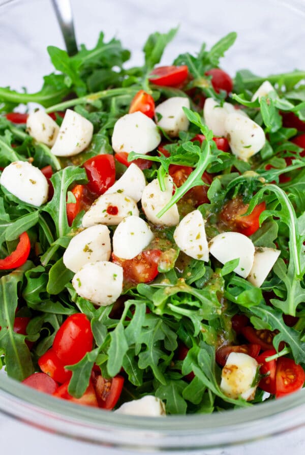 Arugula, tomatoes, and marinated mozzarella balls in glass bowl with spoon.