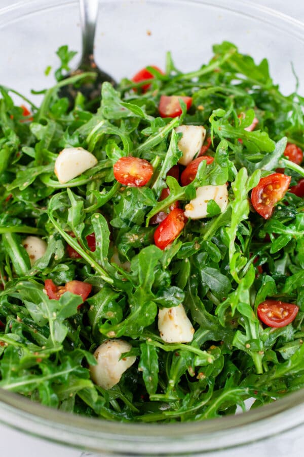 Arugula tomato mozzarella salad tossed in glass bowl with spoon.