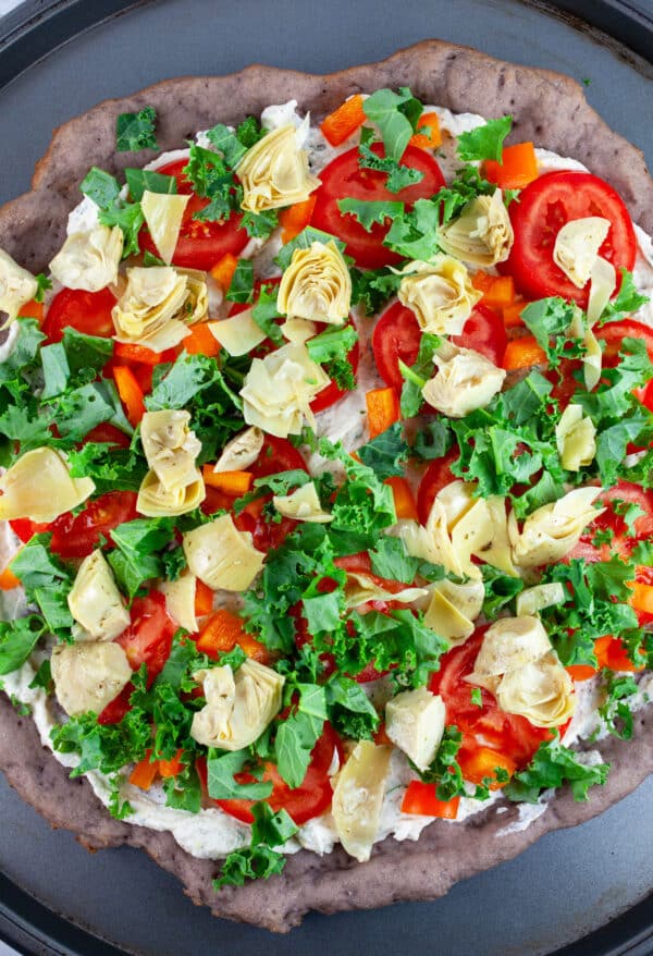Tomatoes, bell peppers, kale, and marinated artichokes on par-baked gluten free pizza crust on pizza pan.