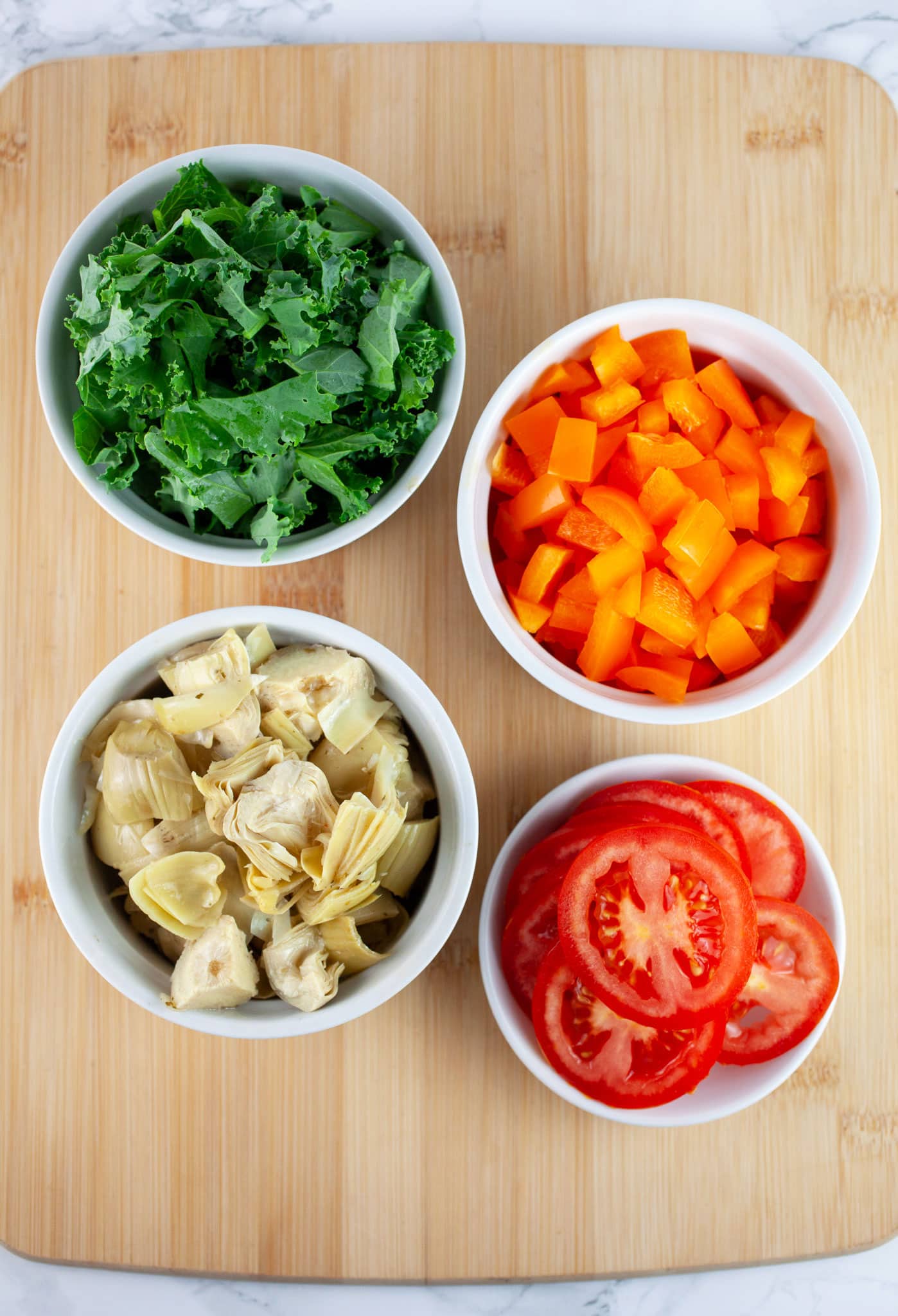 Sliced Roma tomatoes, diced orange bell peppers, chopped kale, and marinated artichoke hearts in small white bowls on wooden cutting board.