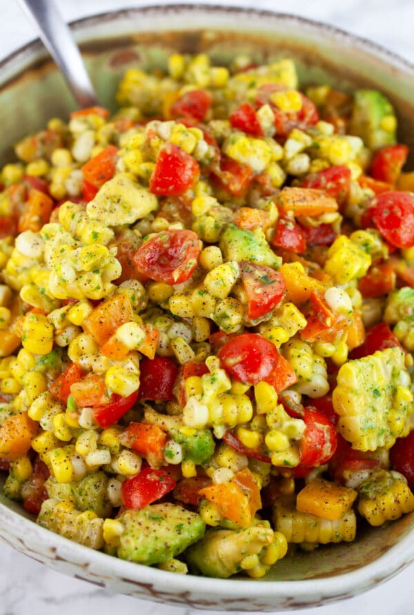 Tomato corn avocado salad with basil vinaigrette in ceramic bowl with metal spoon.