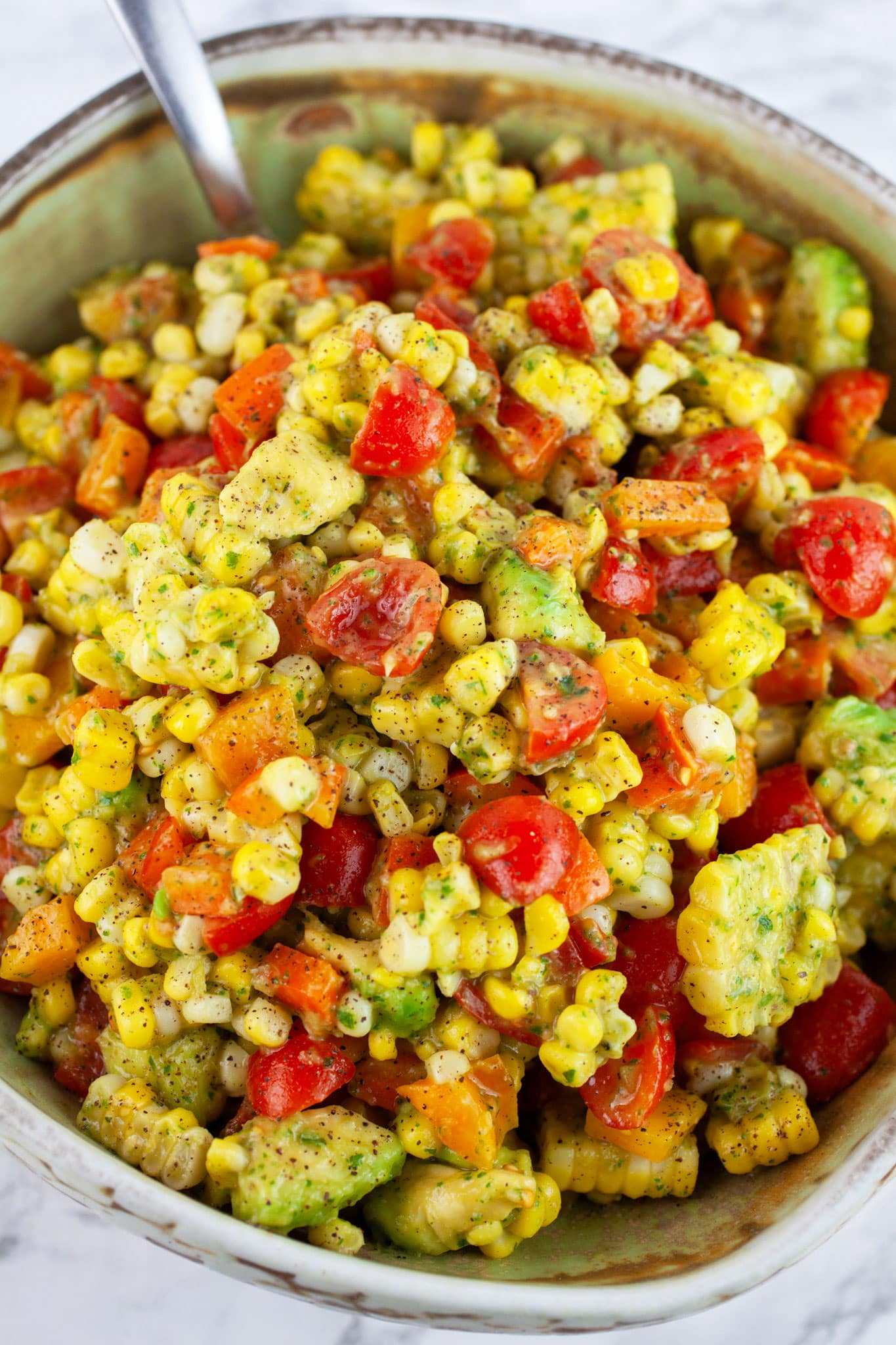 Tomato corn avocado salad with basil vinaigrette in ceramic bowl with metal spoon.