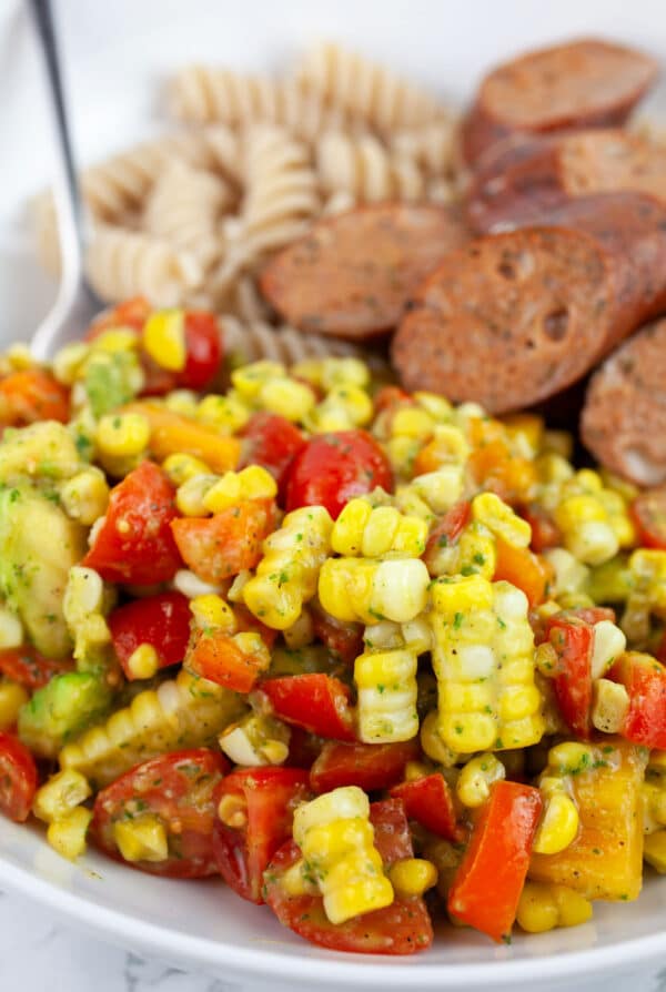 Tomato corn salad in white bowl with fork and chicken sausages and pasta.