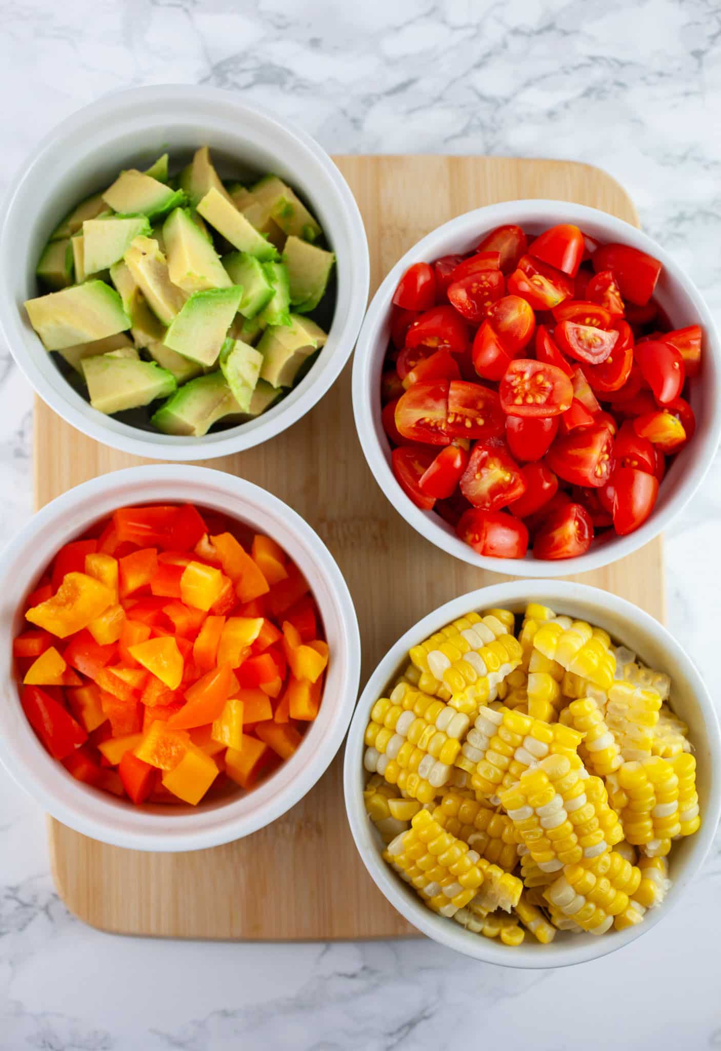 Sweet corn, bell peppers, grape tomatoes, and avocadoes in small white bowls on wooden cutting board.
