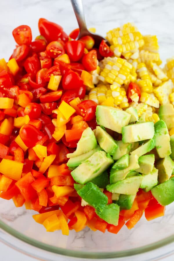 Sweet corn, grape tomatoes, bell peppers, and avocado in large glass bowl with metal spoon.