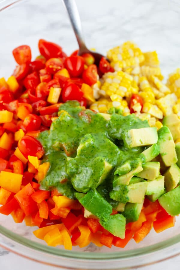 Basil vinaigrette poured onto sweet corn, tomatoes, bell peppers, and avocadoes in large glass bowl with spoon.