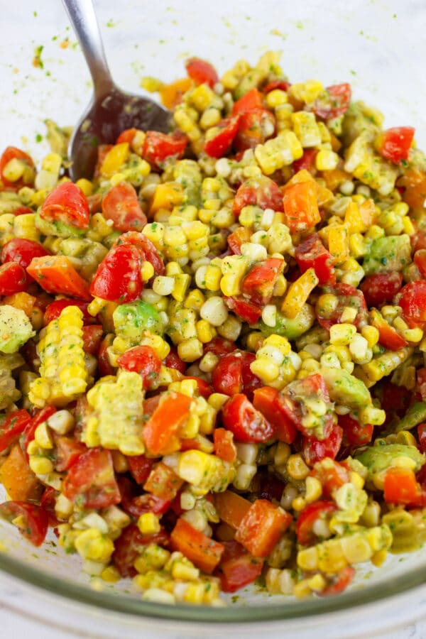 Tomato corn salad with avocadoes and basil vinaigrette in large glass bowl with metal spoon.