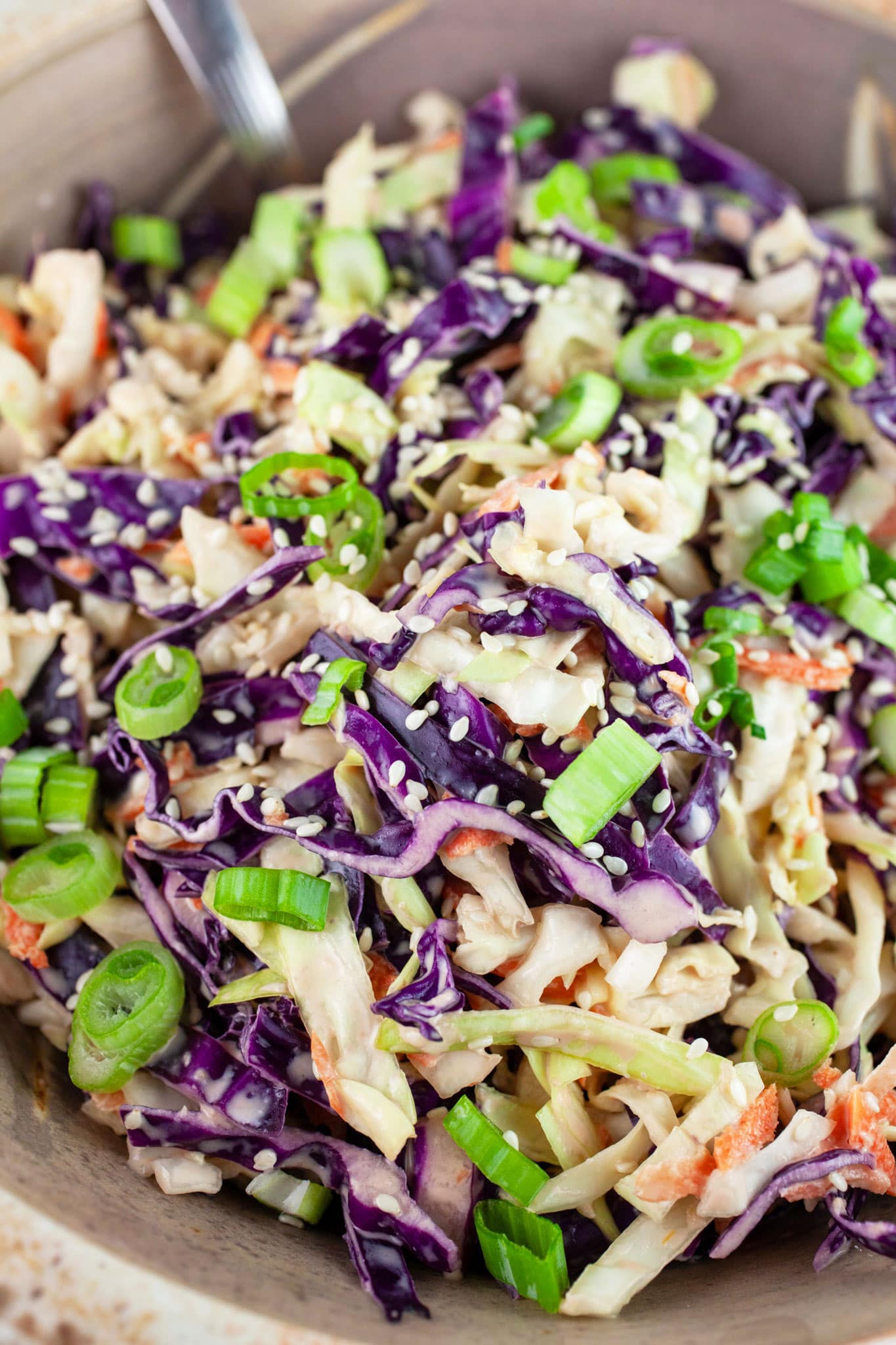 Asian cabbage slaw in ceramic bowl with spoon garnished with green onions and sesame seeds.