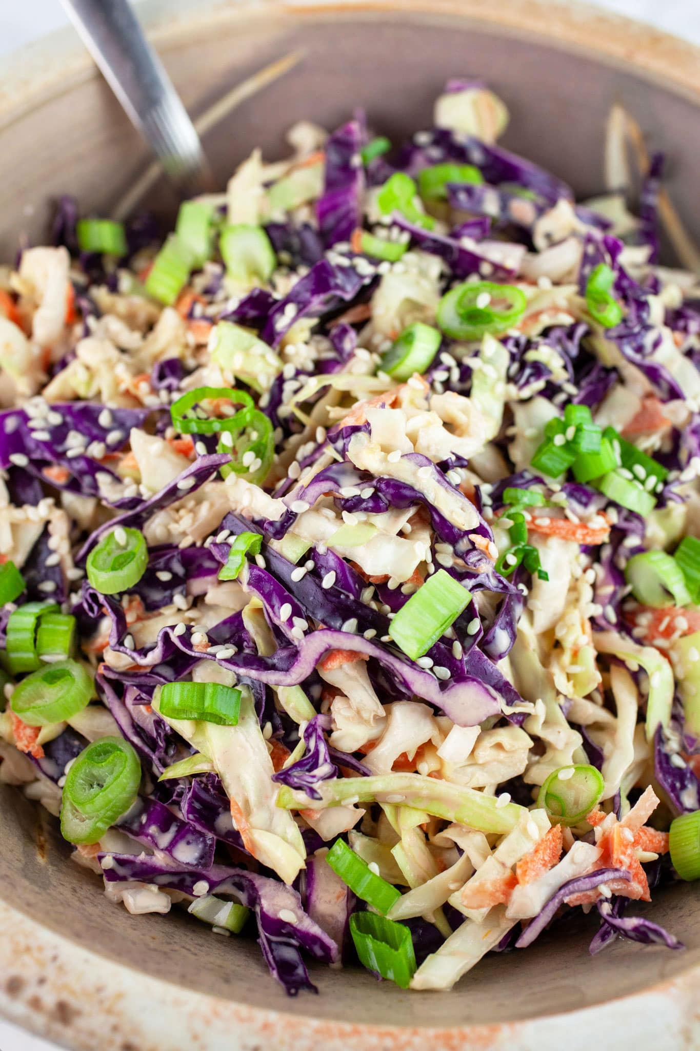 Asian cabbage slaw garnished with green onions and sesame seeds in ceramic bowl with spoon.