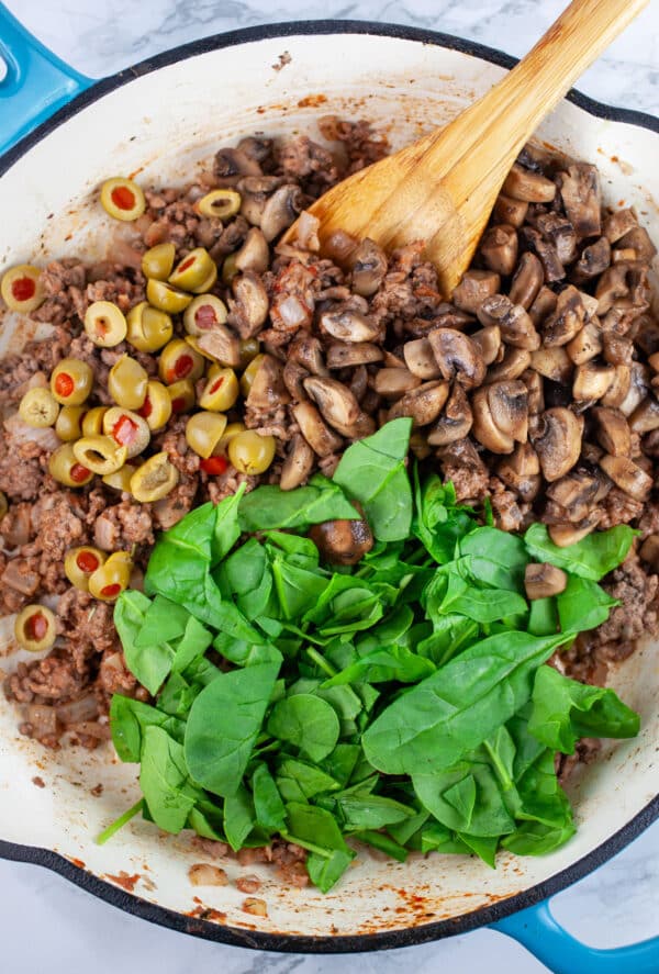Cooked ground beef with spinach and green olives in skillet with wooden spoon.