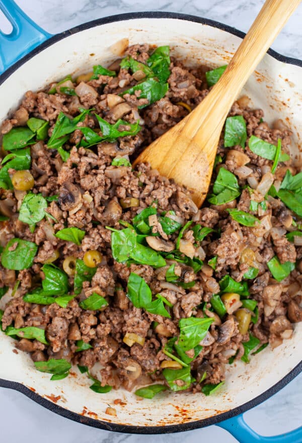 Ground beef, spinach, and green olive mixture in skillet with wooden spoon.