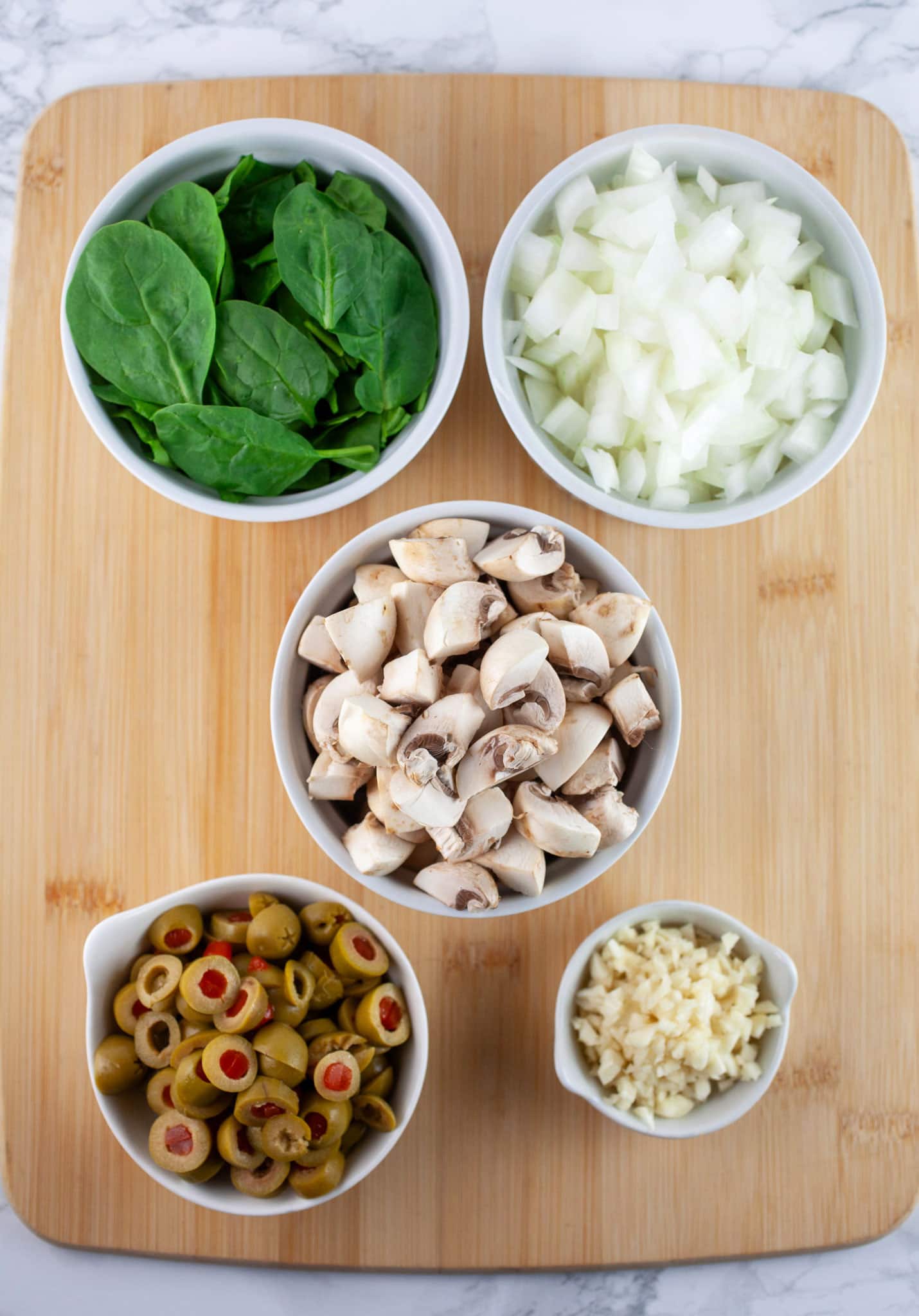 Minced garlic and onions, chopped mushrooms and green olives, and spinach in small white bowls on wooden cutting board.