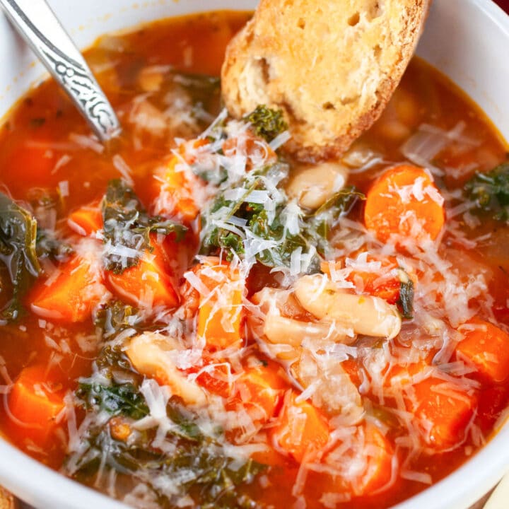 Cannellini bean kale soup with carrots garnished with Parmesan cheese and crostini in white bowl with spoon.