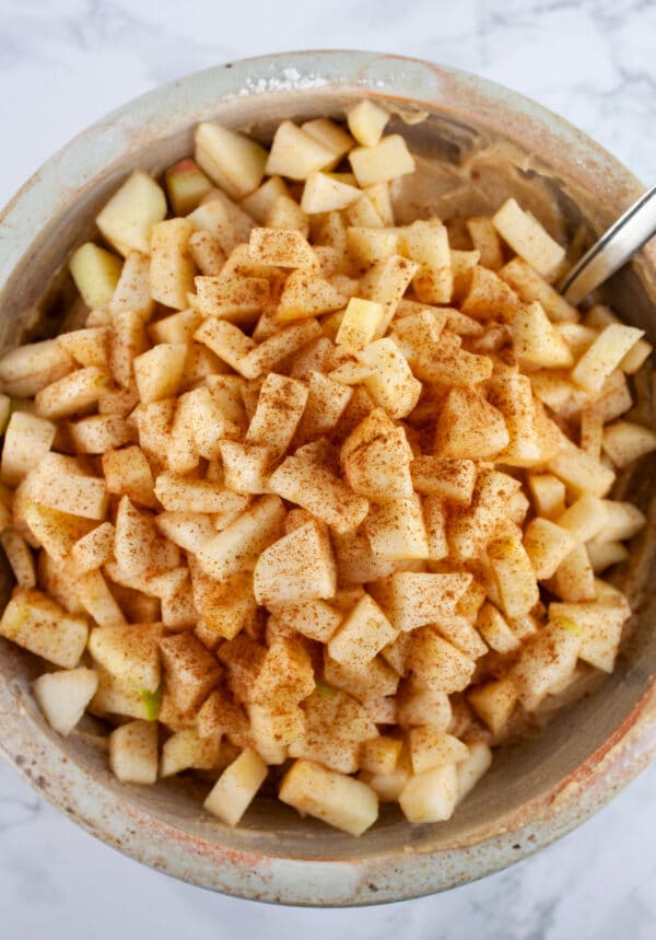 Chopped apples with cinnamon on top of cake batter in ceramic bowl with spoon.