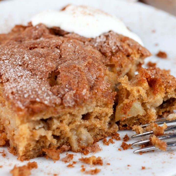 Slice of gluten free apple cake on white plate with fork topped with whipped cream.
