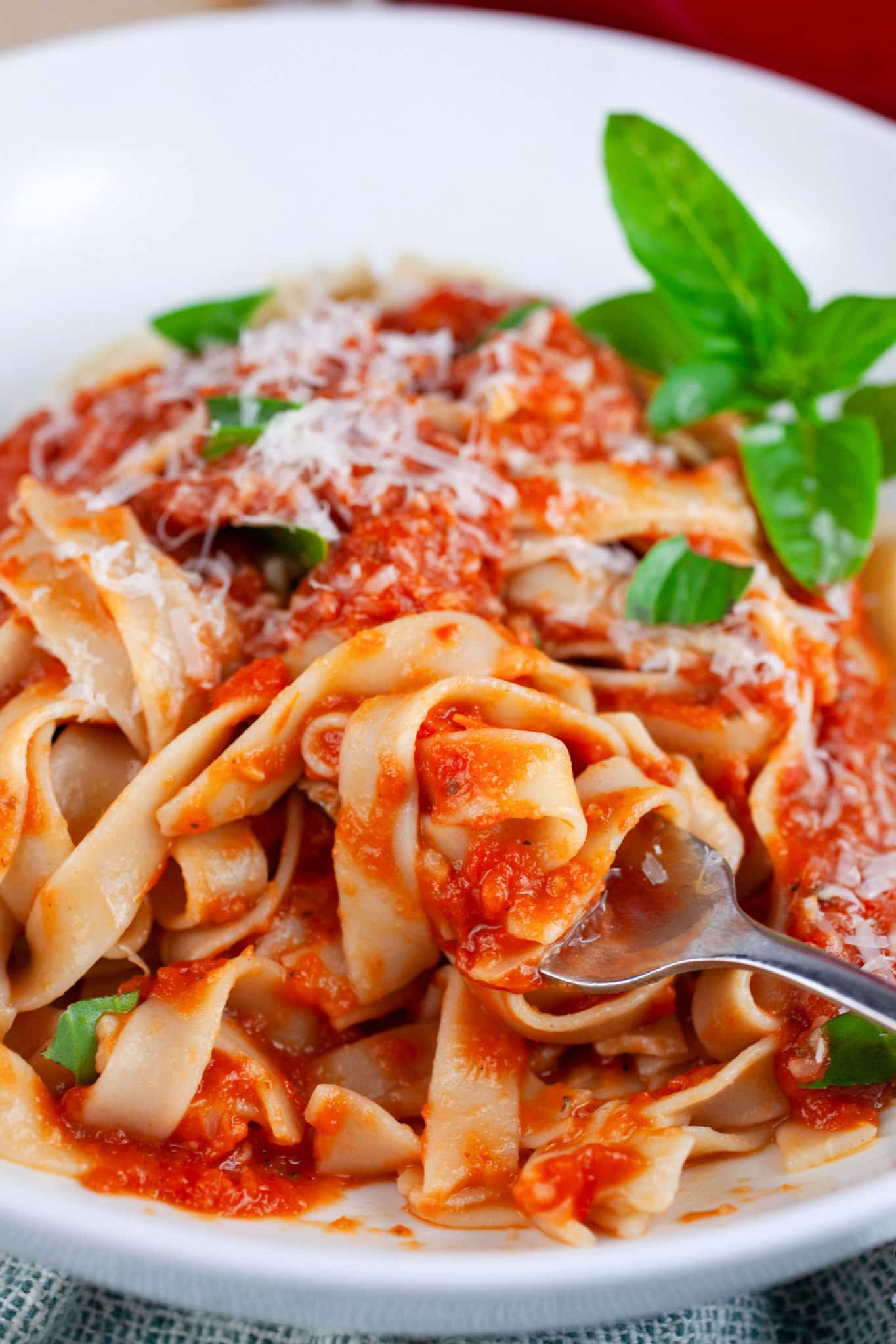 Tagliatelle pasta tossed with roasted tomato sauce in white bowl with fork garnished with Parmesan cheese and fresh basil.