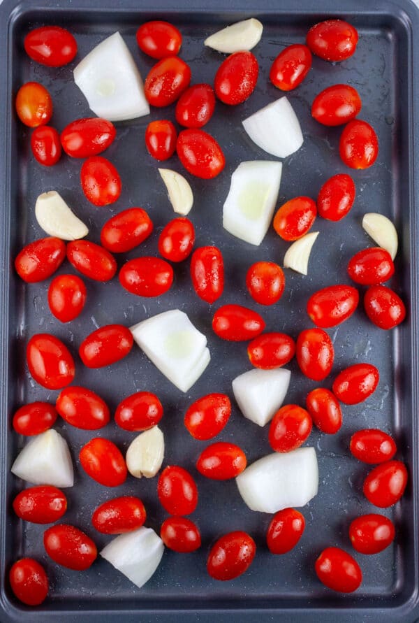 Uncooked grape tomatoes, garlic cloves, and chopped onions on baking sheet tossed in olive oil.