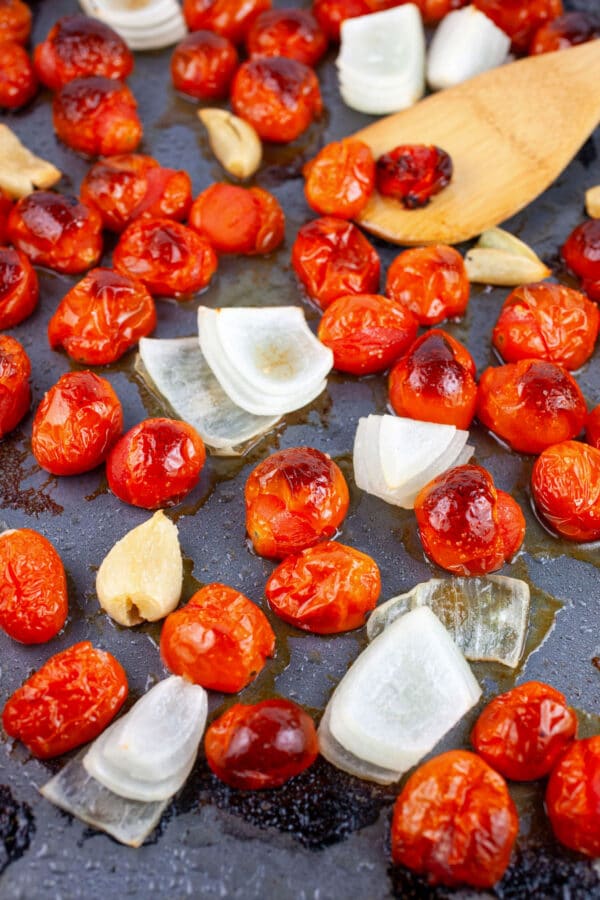 Roasted grape tomatoes, garlic, and onions on baking sheet with wooden spatula.
