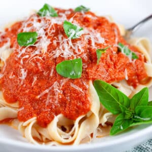 Roasted tomato pasta sauce over tagliatelle in white bowl with fork garnished with Parmesan cheese and fresh basil.